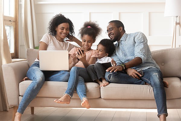 Happy-african-parents-and-kids-obsessed-addicted-to-gadgets-use-laptops-digital-tablet-phone-sit-on-sofa
